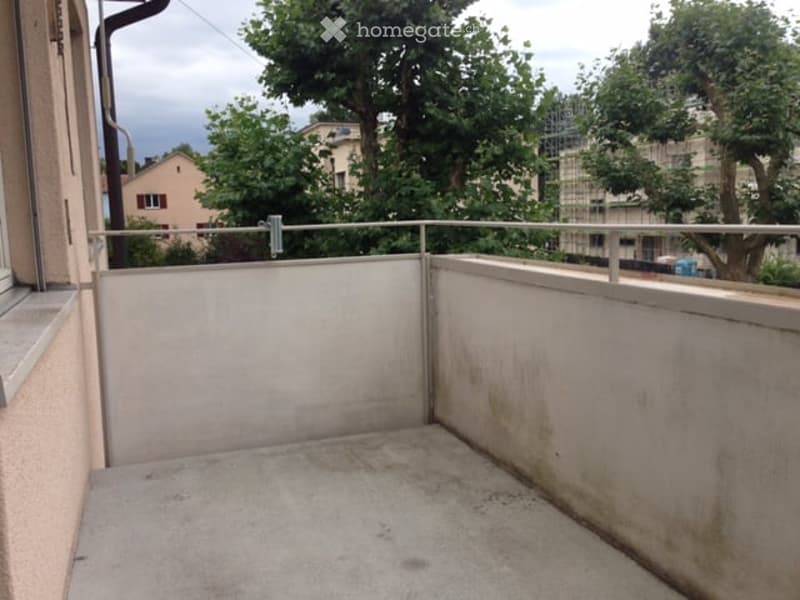 Empty balcony with concrete floor and metal railing, overlooking a residential area with trees and buildings.