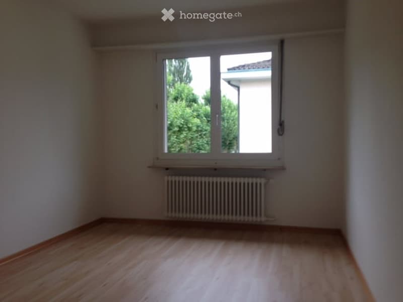Empty room with wooden flooring, white walls, a single window with white frame, and a radiator below the window.