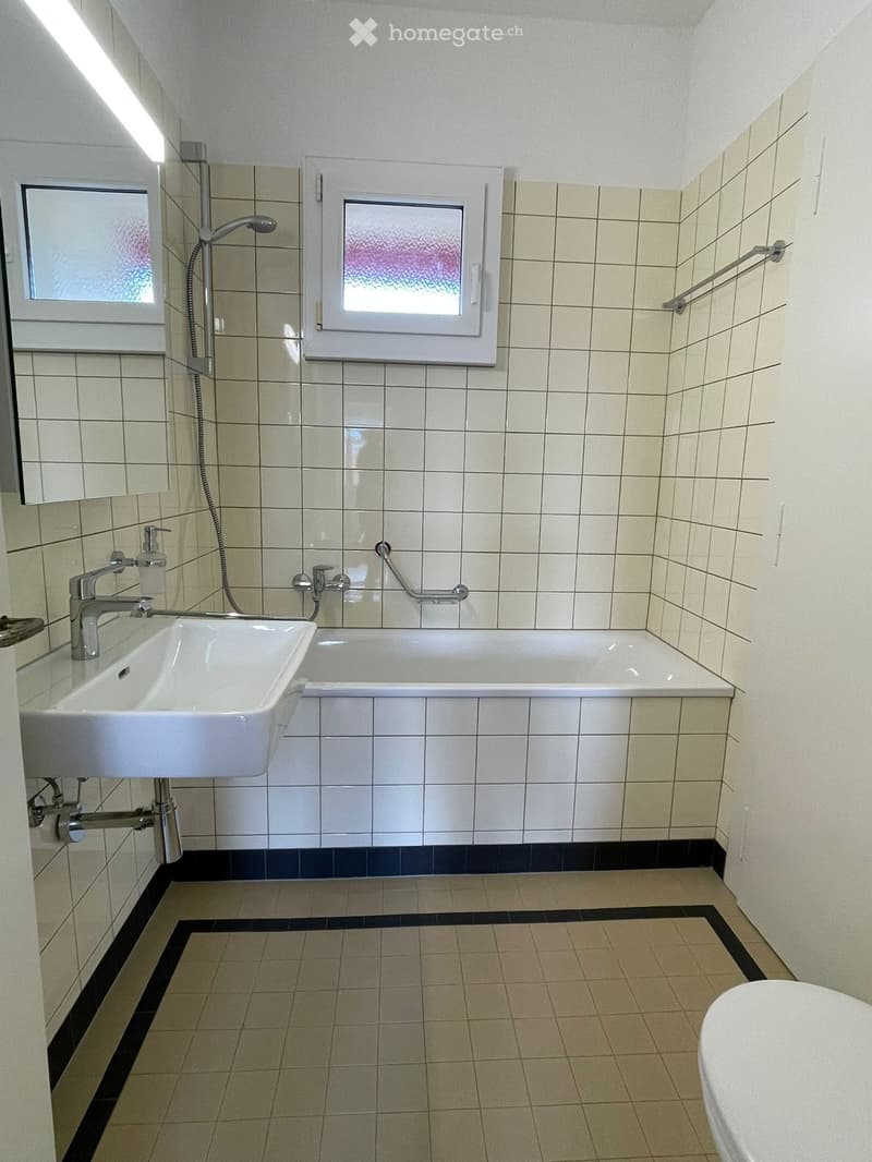 Modern bathroom with a white sink, bathtub, and mirror. Features tiled walls and floor, with a window and handrail for accessibility.