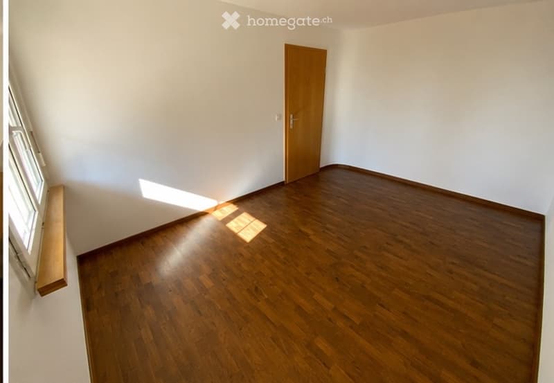 Empty room with wooden flooring, white walls, a single wooden door, and a window allowing natural light to enter.