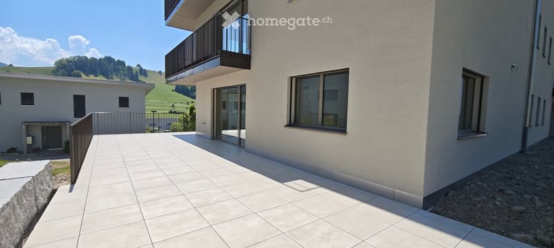 Modern outdoor patio area with white tiled flooring, adjacent to a contemporary building with large windows and a balcony. The background features a scenic green landscape with hills and trees.