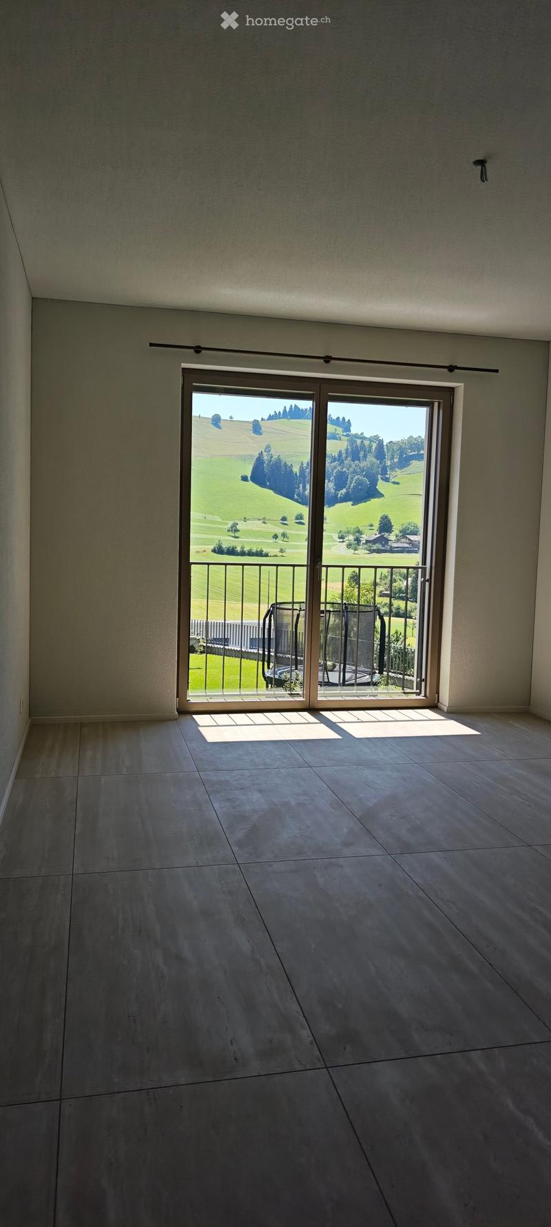 Empty room with large sliding glass doors opening to a balcony, overlooking a green landscape with hills and trees.
