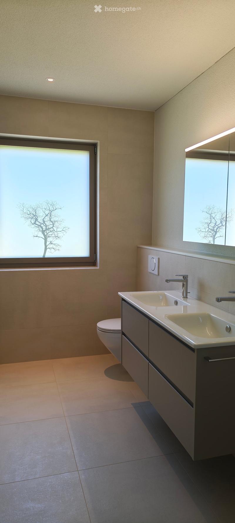 Modern bathroom with a double sink, sleek faucet, large mirror, and a toilet. The room features light-colored tiles and a large window with a tree silhouette design.