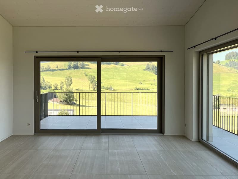 Empty room with large sliding glass doors leading to a balcony, offering a scenic view of green fields and trees. The floor is tiled, and the walls are painted in a neutral tone.