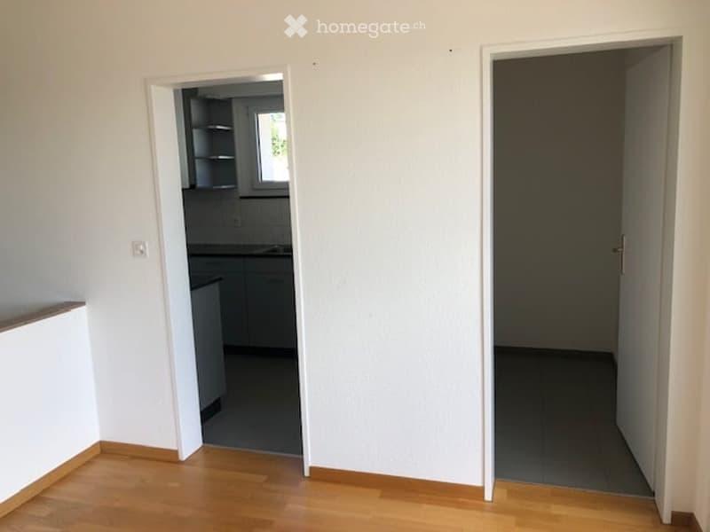 Empty room with white walls, wooden floor, door to the kitchen, and cabinet with shelves