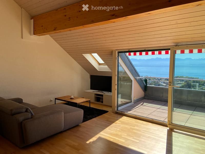 Modern living room with wooden floors, large windows, and a view of a lake and mountains. Features a grey sofa, coffee table, and TV.