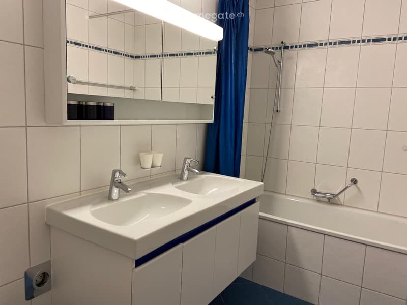 Modern bathroom with white tiles, double sink, mirror, bathtub, and blue shower curtain.