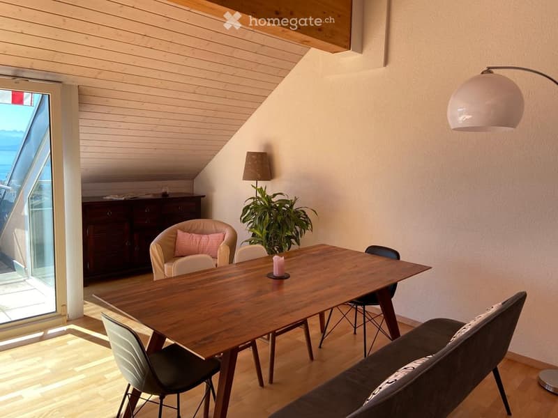 Bright, modern living space with wooden floors, a large window offering a scenic view, a sofa, a dining table with chairs, an armchair, and decorative lamps.