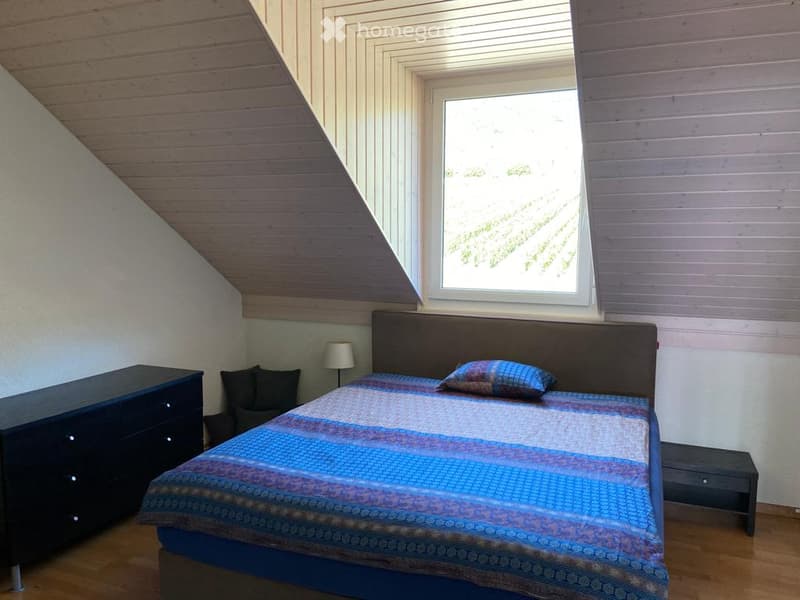 Cozy attic bedroom with wooden slanted ceiling, large window, bed with colorful bedding, nightstand, lamp, and small chair.