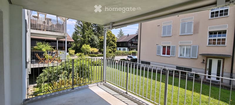 Balcony with metal railing overlooking a green lawn and neighboring buildings, with a view of trees and a parked car.