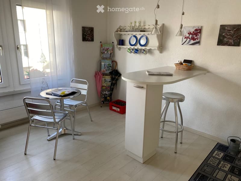 modern kitchen-style room with island counter, 2 chairs, stool, umbrellas, table with a book, white paint, light hardwood floor, small decorative shelf