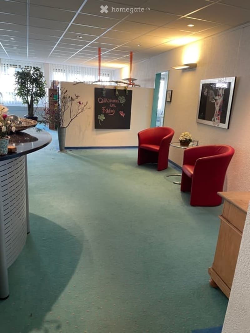 Empty reception area with white walls, two red chairs, small table with flowers, blackboard, framed picture, potted plants, and flowers on a countertop.