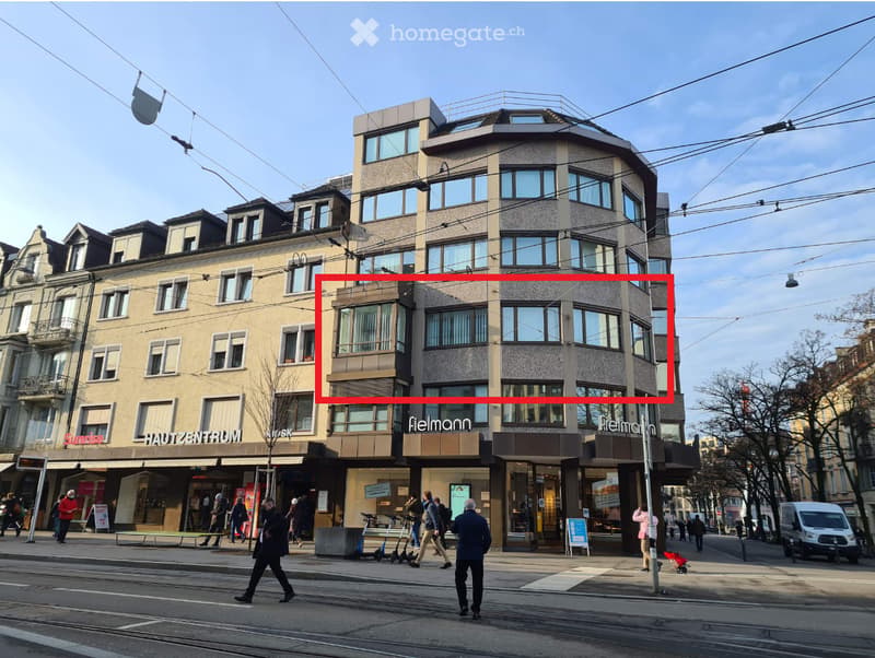 Corner building with several floors, gray exterior, large windows, stores on the ground floor