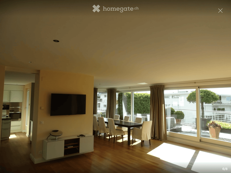 Bright living and dining area with wooden flooring, large windows, a flat-screen TV, a dining table with chairs, and a small cabinet with a stool.