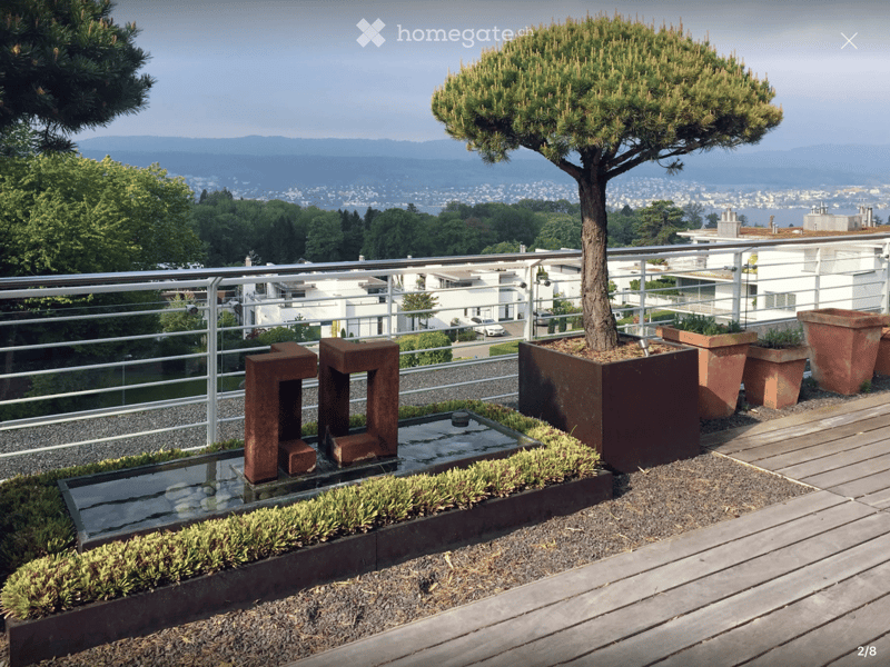 A modern rooftop terrace with a wooden deck, metal railing, decorative water features, potted plants, and a small tree, overlooking a cityscape and mountains.