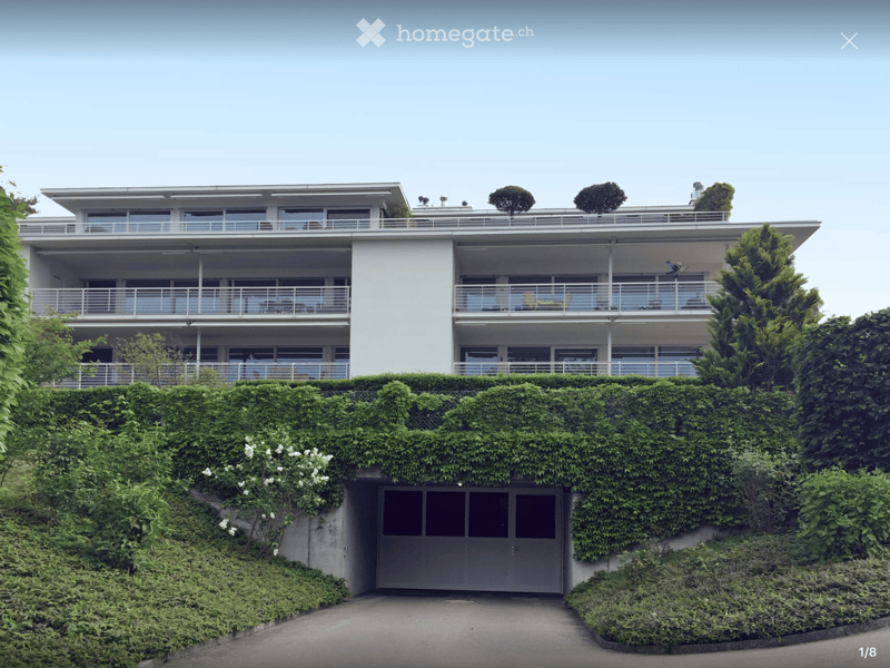 Modern two-story house with white exterior, large windows, balconies, and a garage partially hidden by greenery.