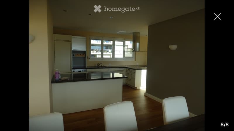 Modern kitchen with white cabinets, a central island, and a dining area with white chairs and a table. The kitchen features a sink, oven, and a bottle on the counter.