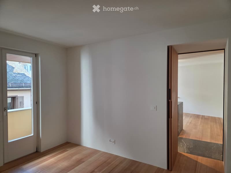 Empty room with light-colored walls and wooden flooring, featuring a door leading to a balcony and an open doorway to another room.