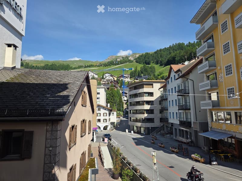 A picturesque village street with multi-story buildings, balconies, and a clear view of green hills and mountains in the background.