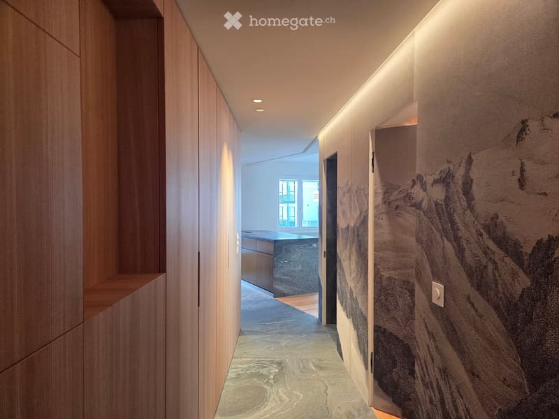 Modern hallway with wooden paneling, recessed lighting, and a textured wall mural. The floor features a polished marble design, and the hallway leads to an open area with a visible kitchen counter and window.