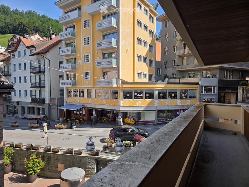 View from a balcony overlooking a town square with shops, restaurants, and a yellow hotel building with balconies. There are potted plants, a parked car, and a bench visible.