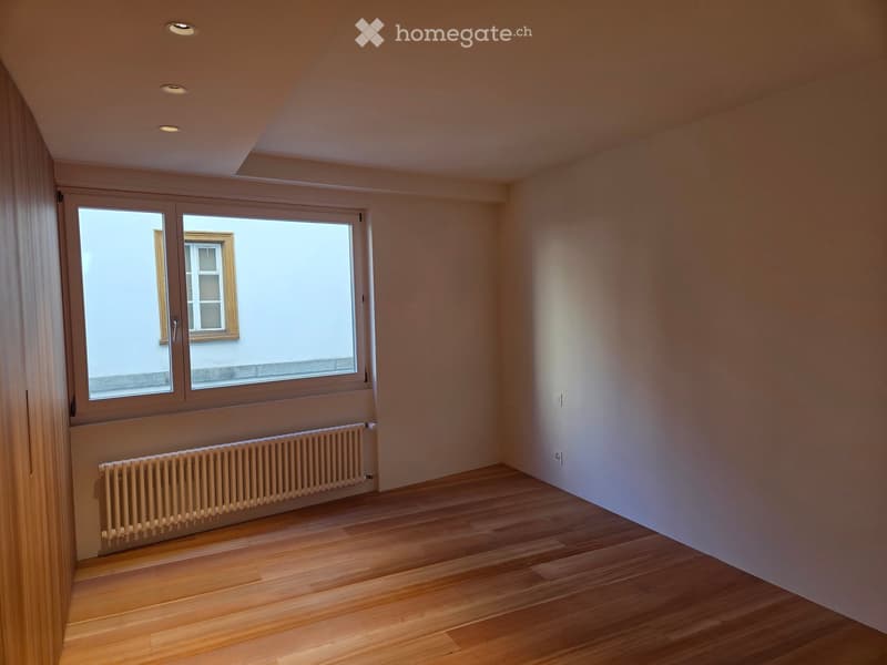 Empty room with wooden flooring, large window, radiator, and recessed ceiling lights.