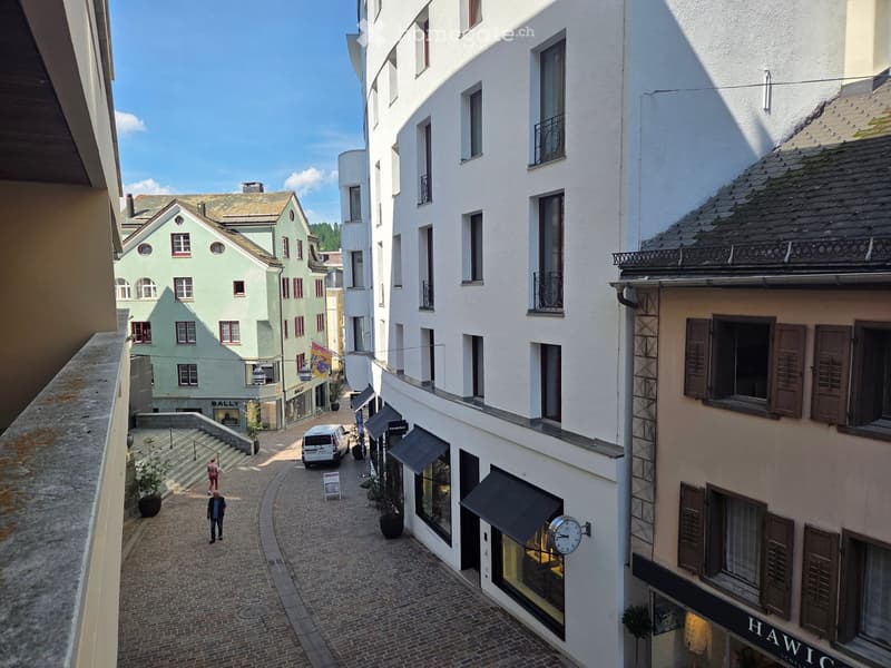 View from a balcony overlooking a narrow cobblestone street lined with multi-story buildings, shops, and pedestrians.