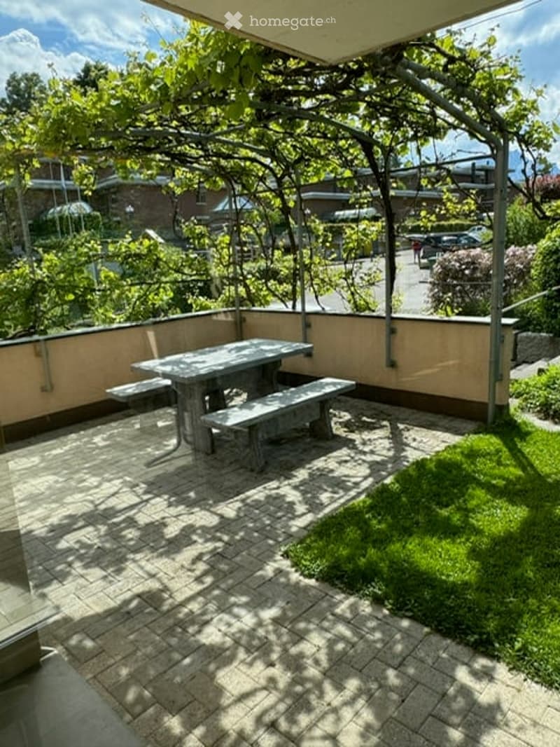 A small outdoor balcony area with a table and benches, surrounded by greenery and a view of the street.