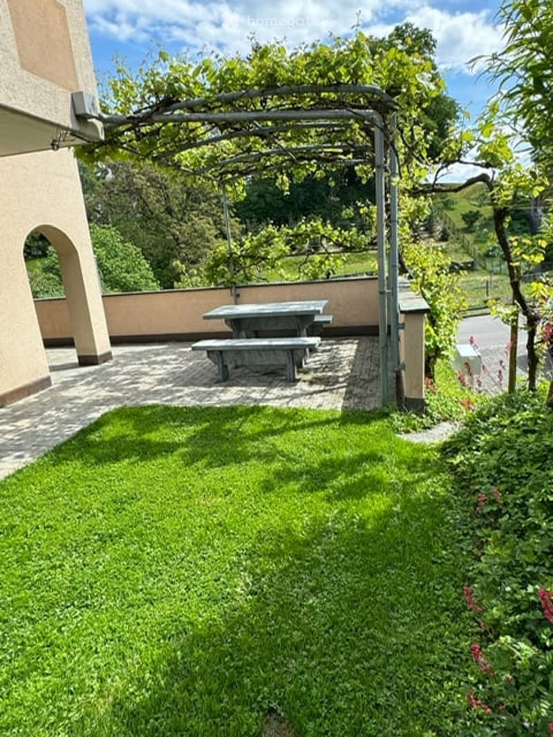 A well-maintained garden area with a green lawn, a covered seating area with picnic tables, and surrounding trees and shrubs.