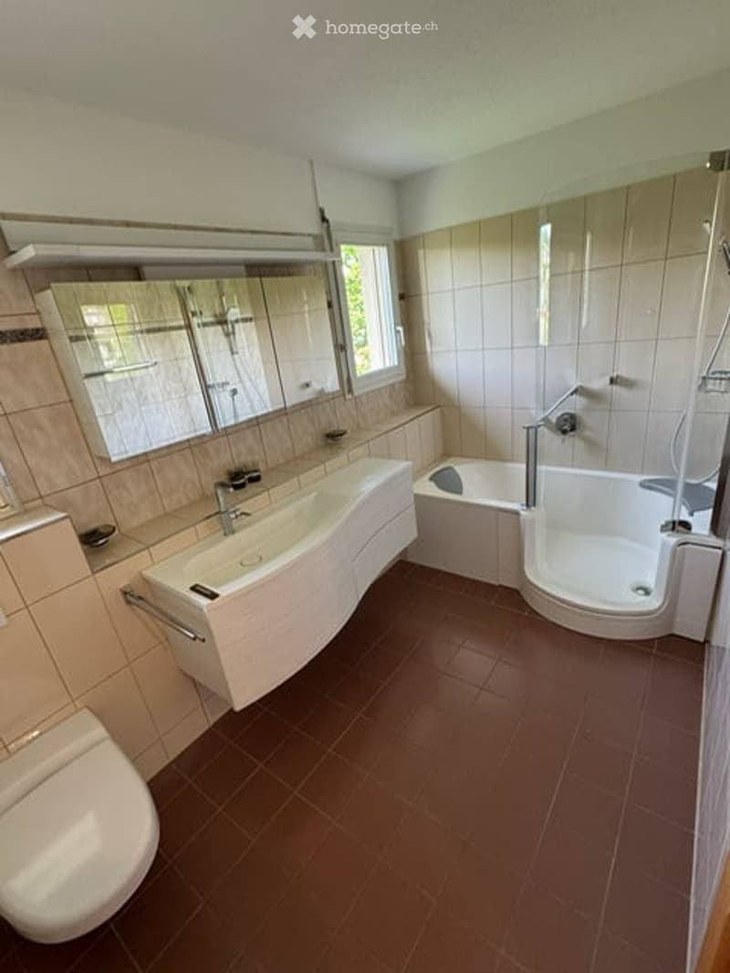 Modern bathroom with a white sink, bathtub, toilet, and large mirror above the sink. The walls are tiled in beige, and the floor is covered with dark brown tiles.