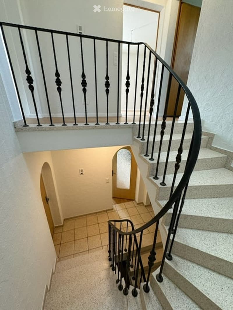 Spiral staircase with black wrought-iron railing and beige tiled landing leading to a lower level hallway with arched doorways.