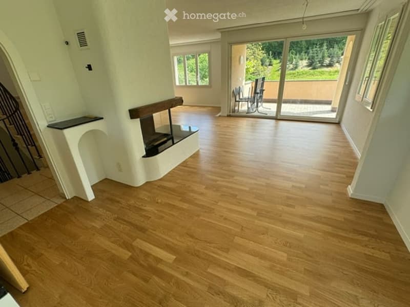 Empty living room with light wooden flooring, white walls, and a fireplace. Large sliding glass doors lead to a patio area with outdoor furniture visible.