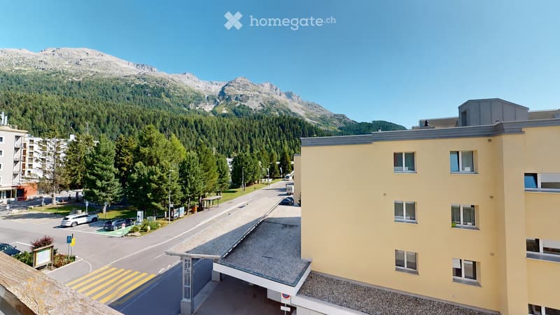 A modern yellow building with multiple windows, situated in a scenic mountain town with a clear view of surrounding peaks and a forested valley.