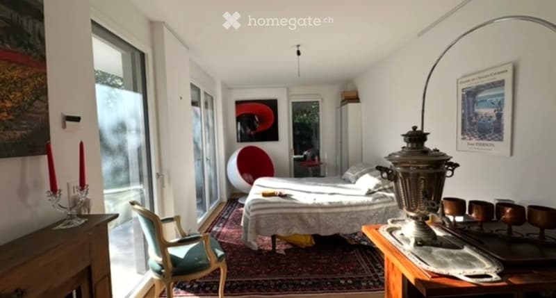 Cozy bedroom with a double bed, a wooden nightstand, a decorative chair, a round red chair, and a large window letting in natural light.
