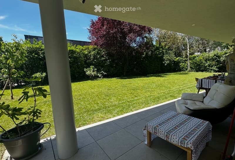 A well-maintained garden with a green lawn, surrounded by trees and shrubs. A patio area features a white leather chair, a small table with a patterned cloth, and a potted plant. The area is covered by a roof supported by white columns.