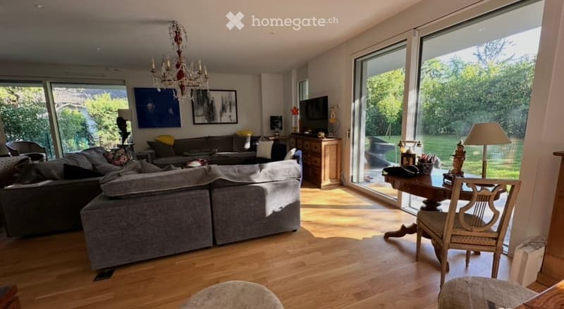 Modern living room with large windows, grey sectional sofa, coffee table, armchairs, ottoman, floor lamp, TV, and a view of the garden.