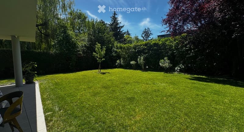 Well-maintained garden with a neatly trimmed lawn, surrounded by trees and shrubs, with a patio area featuring a chair and small table.