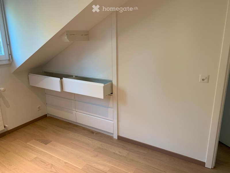 An empty attic space with wooden flooring, white built-in storage units, and a slanted ceiling with a small window.