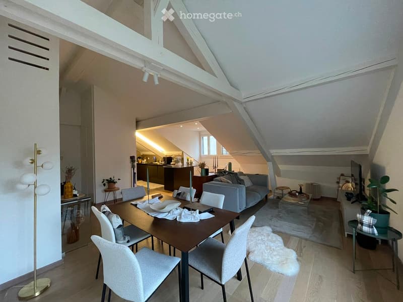 Open-concept living and dining area with a modern aesthetic, featuring a grey sofa, dining table with chairs, floor lamp, and kitchen area visible in the background.