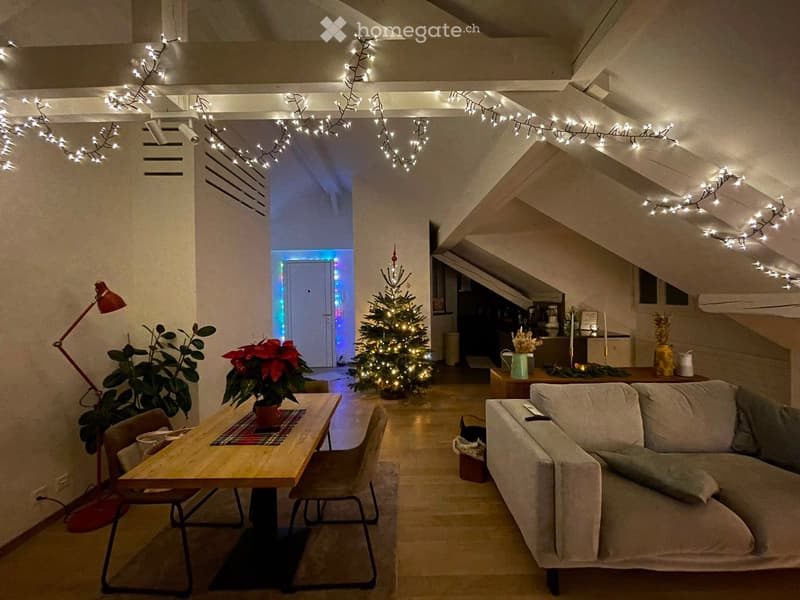Cozy living room with a sofa, coffee table, chairs, lamp, plants, and a decorated Christmas tree. The room features exposed wooden beams and string lights on the ceiling.
