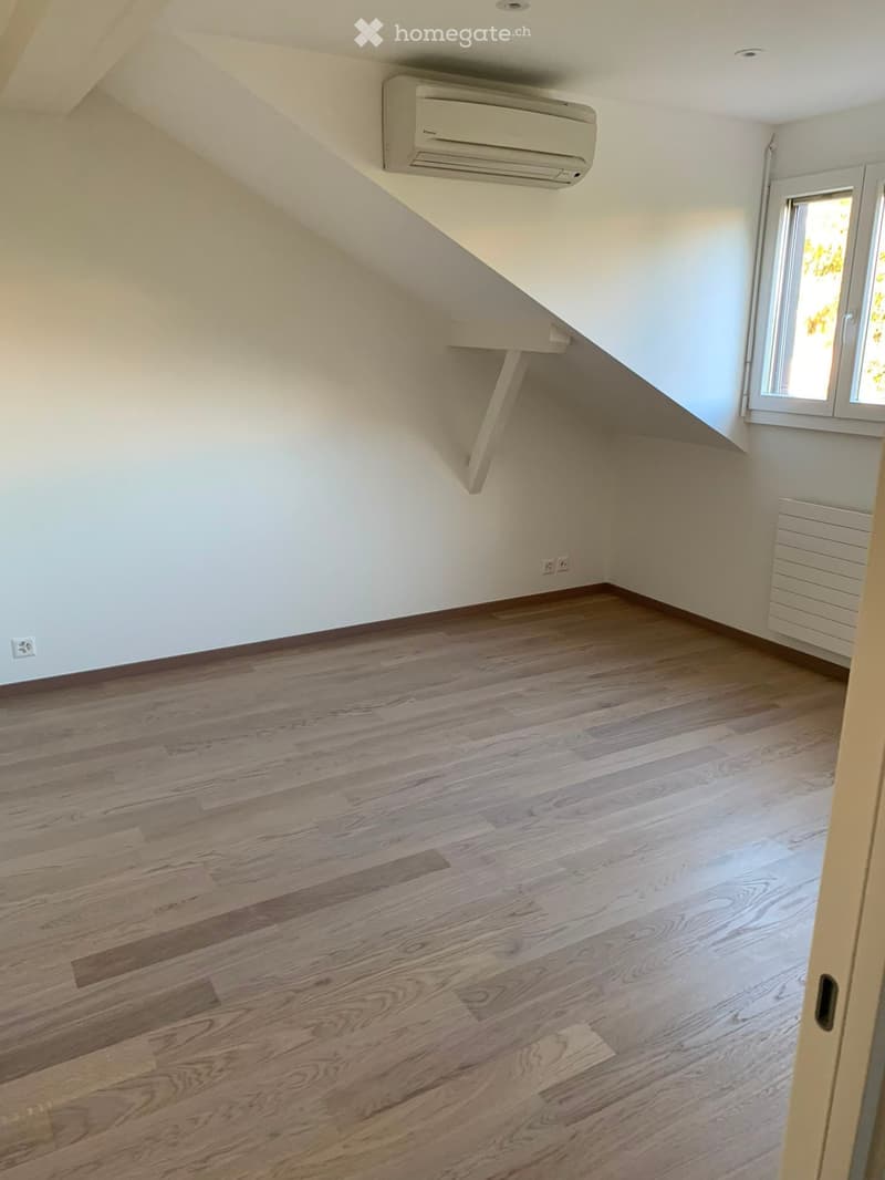 Empty attic room with wooden flooring, white walls, and a window letting in natural light. Features an air conditioning unit mounted on the wall and a radiator on the side.
