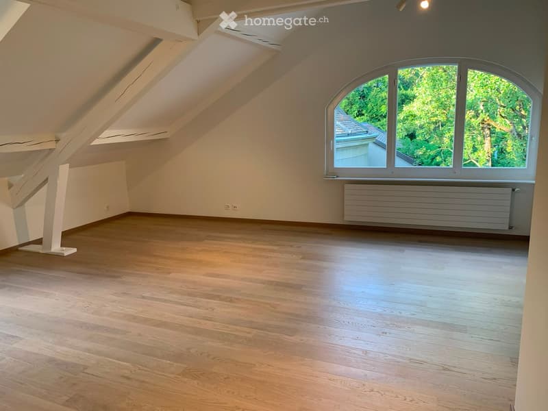 Empty attic room with wooden flooring, slanted ceilings, and a large arched window offering a view of greenery.