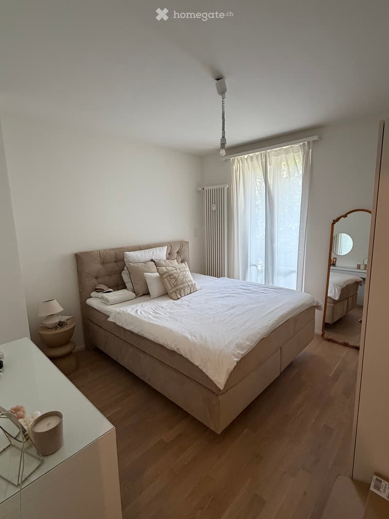Modern bedroom with a large bed, nightstand, lamp, mirror, radiator, and a view through a window with curtains.