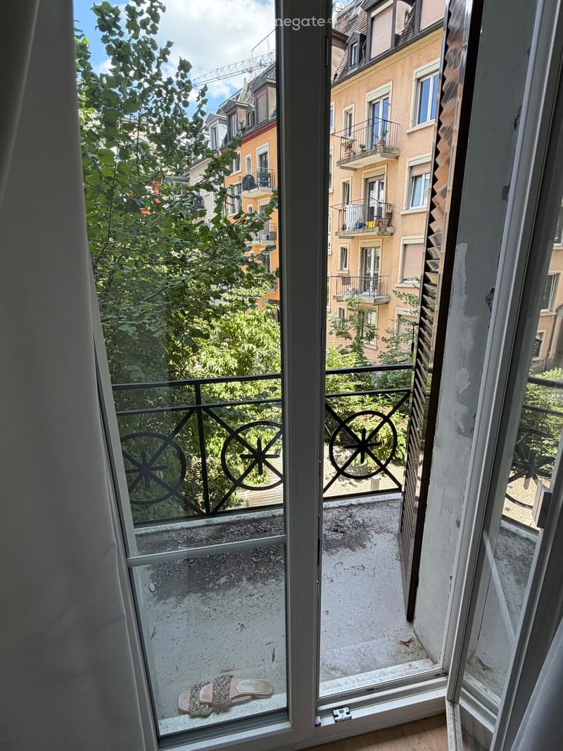 Balcony with metal railing, overlooking a cityscape with trees and neighboring buildings.