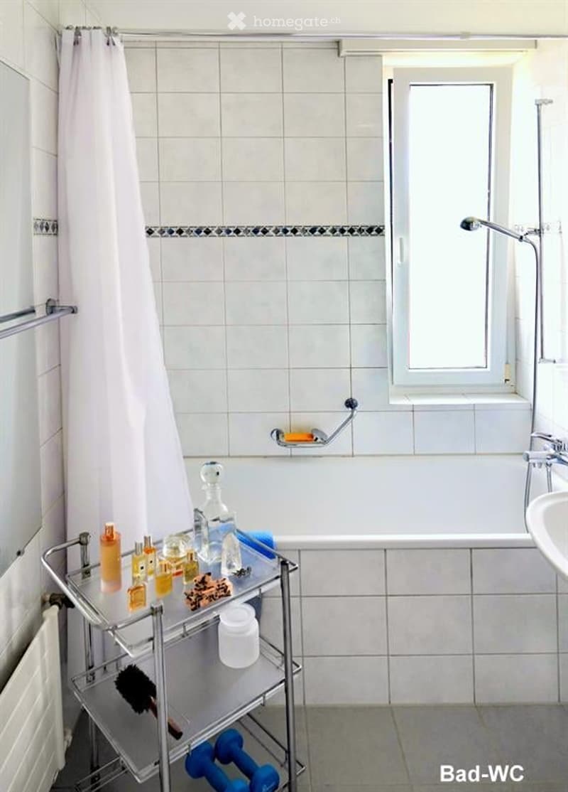 A modern bathroom with a bathtub, shower curtain, and a shelf holding various toiletries and a towel. The room features white tiled walls and a window for natural light.