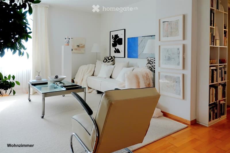 Modern living room with white walls, light-colored carpet, and wooden flooring. Features a white sofa with multiple cushions, a glass coffee table, a single armchair, a bookshelf filled with books, and several decorative picture frames on the walls.