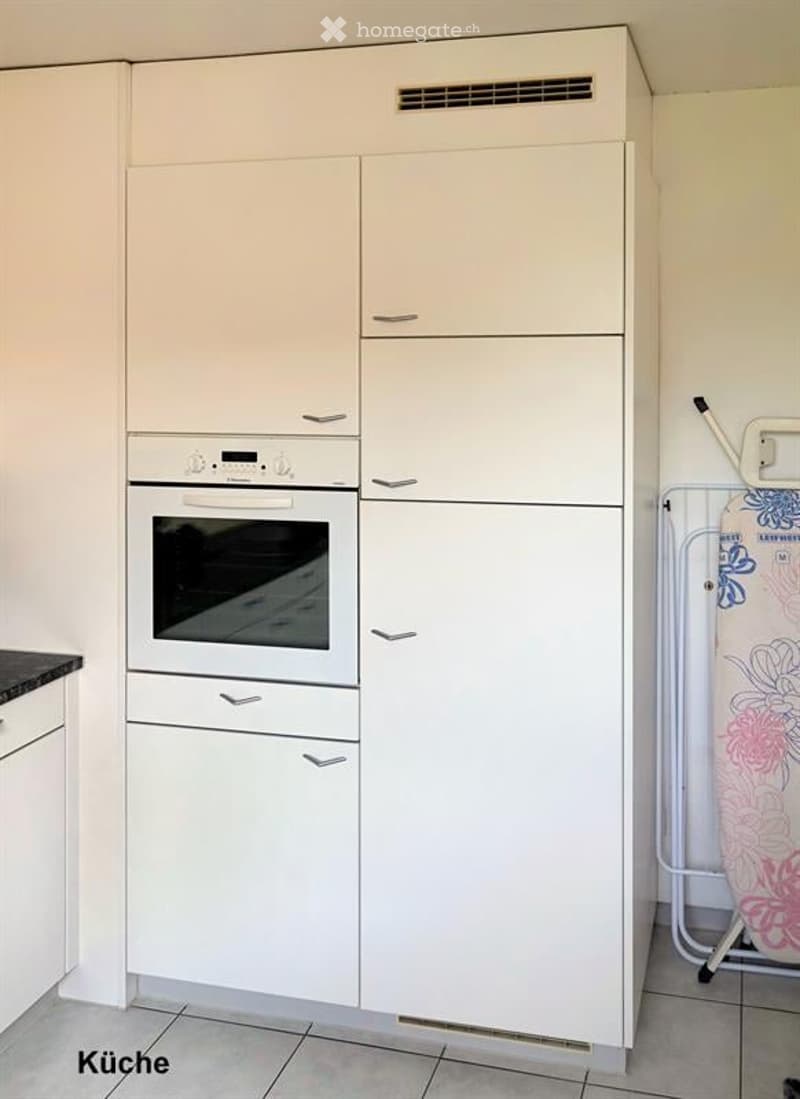 Modern kitchen with white cabinets, an oven, and a refrigerator. A clothes drying rack is visible in the corner.