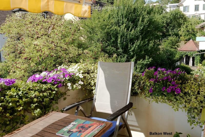 Balcony with wooden table, folding chair, and potted flowers, featuring a yellow and white striped awning and a view of greenery and neighboring houses.