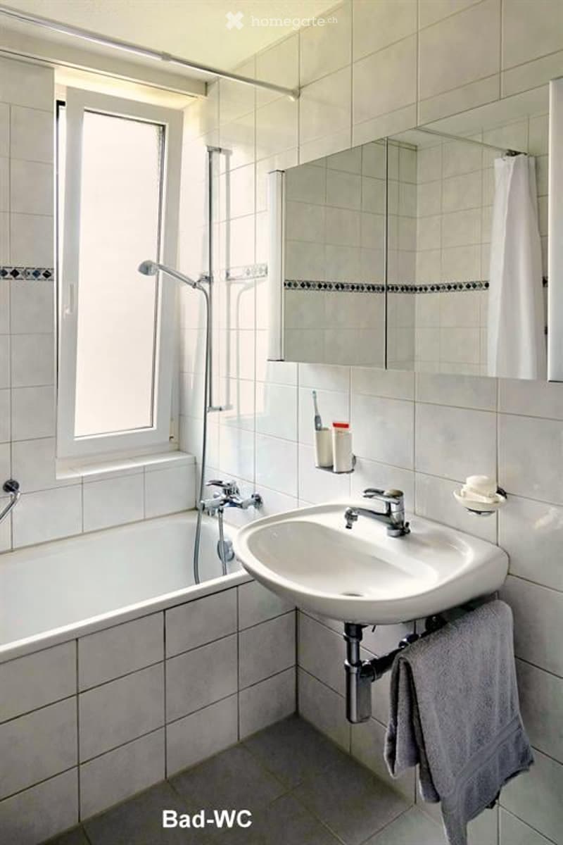 Modern bathroom with white tiled walls and floor, featuring a bathtub, sink, mirror, and towel.