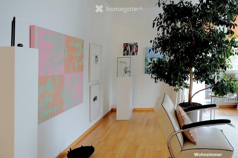 A modern, minimalist living room with light wooden flooring, white walls, and contemporary art pieces. Features include a white armchair with a cushion, a small round table, and a large potted plant near the window.